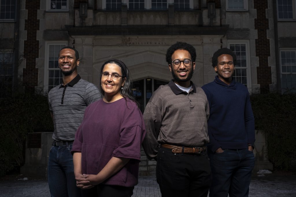 Four members of the UNC Department of Physics and Astronomy -- one professor, two graduate students, and one undergraduate student -- pose for a photo in front of Phillips Hall on UNC's Chapel Hill campus.